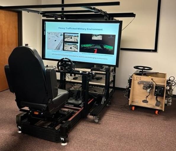 Military vehicle simulation rig showing a driver's seat with steering wheel and a tactical display rendering a heavy trafficked military environment — part of the GVRP digital twin program